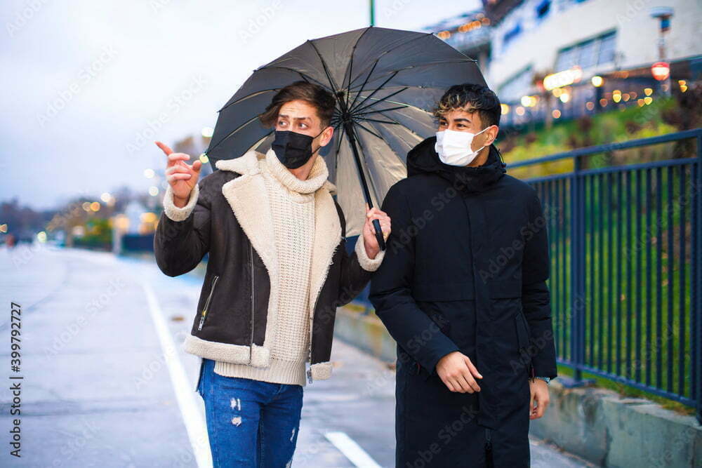 Two friends wearing medical masks on a walk with an umbrella in rainy  weather. Two men